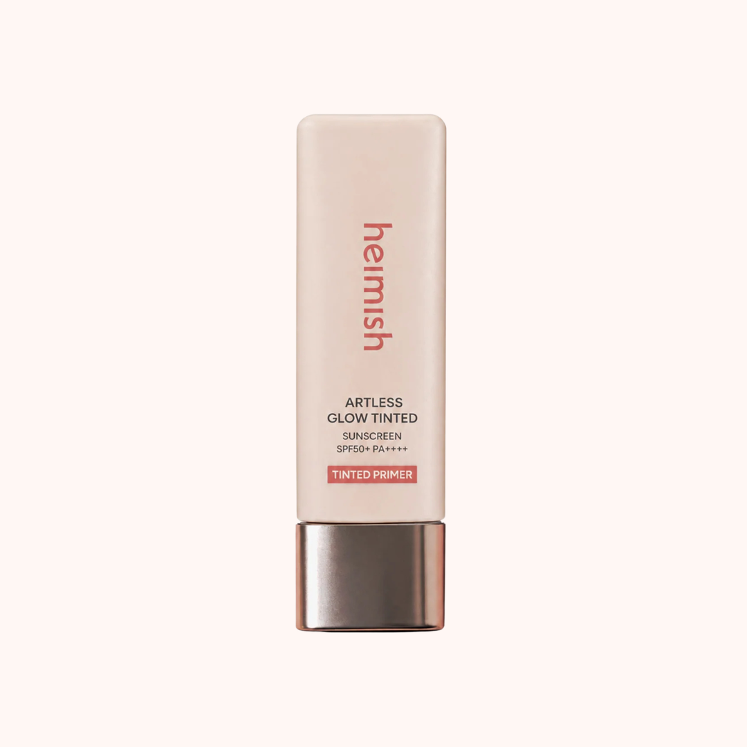 A tube of Hermés Artless Glow Tinted Sunscreen SPF 30 Tinted Primer, featuring a pale pink body and a metallic rose gold cap, centered on a light cream background.